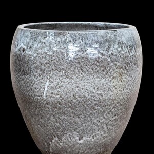 Könnte beinhalten: Eine graue Keramikvase mit strukturierter Oberfläche und breiter Öffnung. Die Vase hat ein gesprenkeltes Aussehen mit weißen Akzenten am Rand. Der Hintergrund ist tiefschwarz.