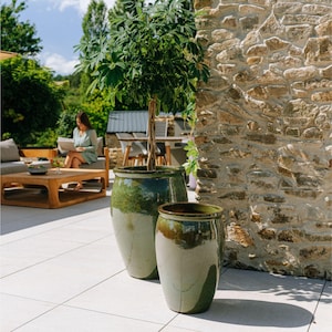 Peut inclure: Deux jardinières en céramique émaillée dans les tons de vert et de crème, avec un arbre dans la plus grande. Les jardinières sont sur un patio carrelé, avec un mur en pierre et des sièges extérieurs en arrière-plan. Les jardinières mesurent environ 60 et 91 cm de haut.