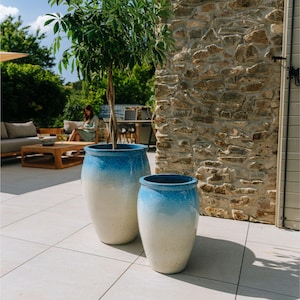 Peut inclure: Deux grands pots de fleurs en céramique fuselés avec une finition lisse et brillante. Les pots présentent un design dégradé, passant d'un bleu profond au niveau du bord à un blanc cassé crème à la base. Le plus grand pot contient un arbre élancé.
