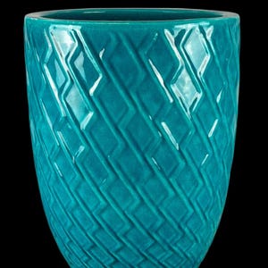 Könnte beinhalten: Eine türkisfarbene Keramikvase mit einem strukturierten Rautenmuster. Die Vase hat eine breite Öffnung und einen abgerundeten Körper. Die glänzende Oberfläche reflektiert das Licht und hebt das komplizierte Design hervor. Die Vase steht vor einem schwarzen Hintergrund.