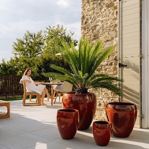 Könnte beinhalten: Eine Sammlung von vier glasierten Terrakotta-Pflanzgefäßen in verschiedenen Größen und einem satten, rotbraunen Farbton. Die Pflanzgefäße stehen auf einer hellen Terrasse, mit einer großen Palmpflanze im größten Topf. Im Hintergrund sitzt eine Frau an einem Tisch.