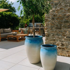 Könnte beinhalten: Zwei große, blau-weiß schattierte Keramik-Pflanzgefäße mit einem Baum. Die Pflanzgefäße stehen auf einer hell gefliesten Terrasse. Im Hintergrund sitzt eine Frau. Die Szene spielt sich im Freien mit einer Steinmauer ab.