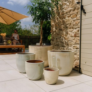 Könnte beinhalten: Eine Sammlung von Keramik-Pflanzgefäßen in verschiedenen Größen und Farben, darunter Beige, Hellgrün und Off-White. Die Pflanzgefäße haben eine glänzende Oberfläche und sind auf einer gefliesten Terrasse angeordnet. Ein Baum ist in einem der größeren Pflanzgefäße gepflanzt.