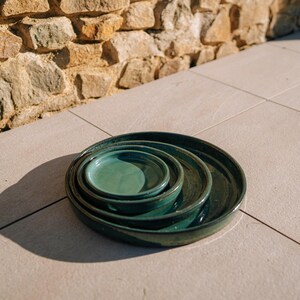 Peut inclure: Un ensemble de quatre assiettes en céramique de couleur teal, de tailles différentes, empilées les unes dans les autres. Les assiettes ont une finition brillante et un bord légèrement surélevé. Elles sont disposées sur une surface carrelée de couleur claire.