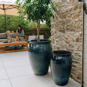 Peut inclure: Deux grands pots de fleurs en céramique arrondis avec une finition brillante et mouchetée bleu profond et sarcelle. Le plus grand pot contient un petit arbre au tronc tressé, tandis que le plus petit est vide sur le sol de la terrasse.