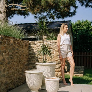 Könnte beinhalten: Drei weiße Keramik-Pflanzgefäße mit einem strukturierten, wirbelnden Design. Die Pflanzgefäße sind unterschiedlich groß und stehen auf einer hellen Terrasse. Eine Frau steht in der Nähe in einem weißen Oberteil und Shorts.