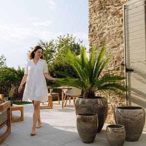 Könnte beinhalten: Eine Frau in einem weißen Kleid geht an einem Set aus vier dekorativen, strukturierten, graubraunen Keramikpflanzgefäßen vorbei. Die Pflanzgefäße sind unterschiedlich groß, wobei ein großes eine grüne Pflanze enthält. Die Szene spielt sich im Freien auf einer Terrasse ab.