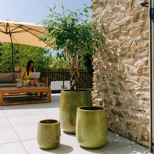 Peut inclure: Trois jardinières en céramique vertes de différentes tailles, avec une finition texturée, sont posées sur un patio. Un petit arbre aux feuilles vertes est planté dans la plus grande jardinière. Une femme est assise en arrière-plan.