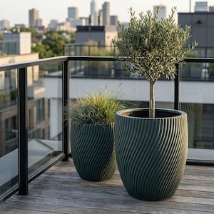 Könnte beinhalten: Zwei große, dunkelgrüne Pflanzgefäße mit Spiraltextur auf einer Holzterrasse, jeweils mit Grünpflanzen. Ein Pflanzgefäß enthält einen kleinen Olivenbaum, das andere Ziergras. Im Hintergrund ist eine Stadtlandschaft zu sehen.