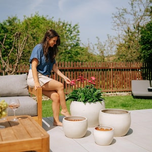 Peut inclure: Ensemble de quatre pots de fleurs en céramique blanche de différentes tailles, de forme arrondie et à surface texturée. Ils sont disposés sur un patio gris clair, avec une personne et un jardin en arrière-plan.