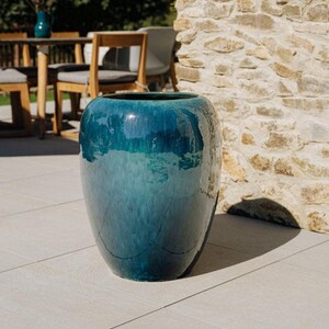Peut inclure: Un grand pot de fleurs en céramique de couleur teal avec une finition brillante. Le pot d'extérieur a une forme arrondie et est placé sur un patio en pierre de couleur claire. Le pot est un élément décoratif pour les jardins et les patios.