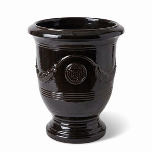 Peut inclure: Un pot de fleurs en céramique vernissée brun foncé avec un bord évasé et une base sur pied. L'extérieur présente un médaillon circulaire en relief avec un emblème central et des guirlandes décoratives sur le corps supérieur.