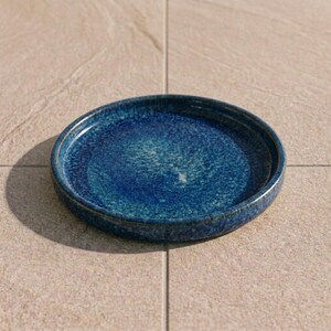 Peut inclure: Une assiette circulaire en céramique dotée d'un vernis bleu profond avec des taches bleu clair et sarcelle. L'assiette présente un bord légèrement surélevé et une finition lisse et brillante, révélant une riche variation de couleurs.