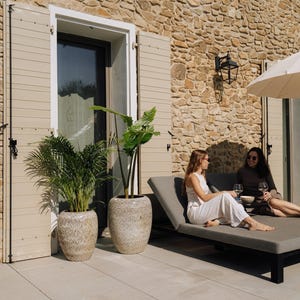 Peut inclure: Deux femmes se détendent sur une chaise longue grise sur un patio, tenant des verres à vin. Le patio comprend de grands pots en pierre avec des plantes vertes. L'arrière-plan est un mur en pierre et des volets beiges.