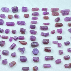 Peut inclure: Une collection de pierres précieuses rubis brutes et non taillées, de formes et de tailles variées. Les pierres varient du rose foncé au violet rougeâtre, certaines présentant des teintes mixtes. Les pierres sont dispersées sur une surface blanche.