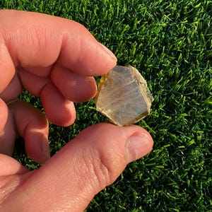 May include: A translucent, light-yellow crystal with a geometric shape is held between two fingers against a backdrop of green artificial turf. The crystal has a multifaceted surface and appears to be naturally formed.