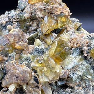 May include: Close-up of a mineral specimen featuring clusters of translucent, golden-yellow crystals. The crystals are set against a matrix of gray, brown, and black rock, creating a striking contrast. The overall texture is rough and uneven.