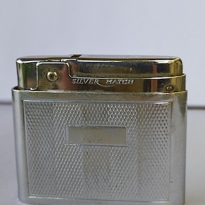 Pode incluir: Um isqueiro prateado com o texto "SILVER MATCH" na parte superior. O isqueiro tem formato retangular com um design texturizado em padrão de diamante na frente e nas laterais. A parte superior tem um acabamento metálico brilhante.