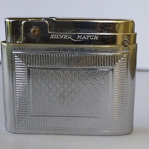 Puede incluir: Un encendedor plateado con el texto "SILVER MATCH" en la parte superior. El encendedor tiene forma rectangular con un diseño texturizado en la parte delantera. El encendedor es un objeto de colección vintage.
