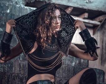May include: A woman wearing a black hooded top with a lace design, a black top with gold chains, a black skirt with fringe, black fishnet stockings, and black boots. She is sitting on a set of concrete steps.
