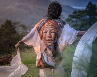 Puede incluir: Una mujer lleva un kimono blanco con un retrato colorido de una mujer en la espalda. El kimono ondea con el viento mientras ella se encuentra en una colina cubierta de hierba con vistas a una cordillera.