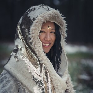 May include: A woman wearing a cream and light gold hooded scarf with a sequined design. The scarf has a faux fur trim.