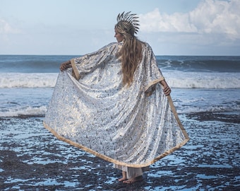 May include: A woman wearing a silver sequined cape with a gold trim stands on a rocky beach with the ocean in the background. She is wearing a gold crown and has long blonde hair.