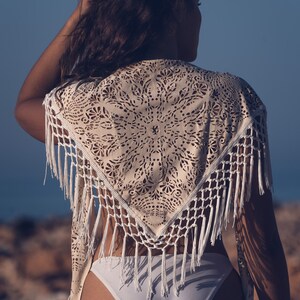 May include: A white crochet shawl with a delicate lace pattern and fringe trim. The shawl is worn over a white bikini bottom.