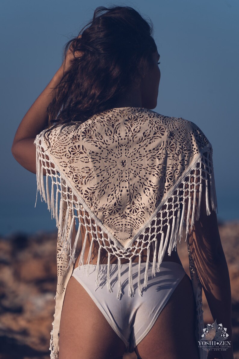 May include: A white crochet shawl with a delicate lace pattern and fringe trim. The shawl is worn over a white bikini bottom.