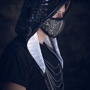 May include: A person wearing a black hooded jacket with a white patterned lining and a black face mask with a geometric design. The person is wearing a silver chain necklace.