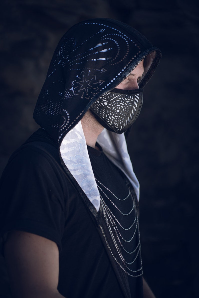 May include: A person wearing a black hooded jacket with a white patterned lining and a black face mask with a geometric design. The person is wearing a silver chain necklace.