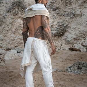 K&ouml;nnte beinhalten: Ein Mann tr&auml;gt eine wei&szlig;e Hose mit einem dekorativen Besatz und einen wei&szlig;en Schal mit einem goldenen Besatz. Er steht an einem Sandstrand mit einer felsigen Klippe im Hintergrund.