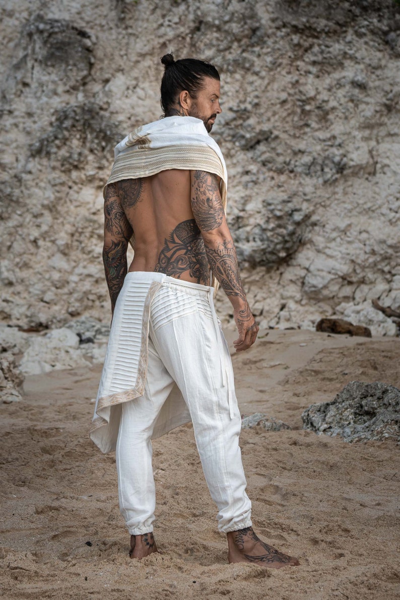 K&ouml;nnte beinhalten: Ein Mann tr&auml;gt eine wei&szlig;e Hose mit einem dekorativen Besatz und einen wei&szlig;en Schal mit einem goldenen Besatz. Er steht an einem Sandstrand mit einer felsigen Klippe im Hintergrund.