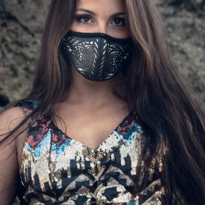 May include: A woman with long brown hair wearing a black face mask with a geometric design. She is wearing a sequined top with a geometric pattern in shades of blue, red, and gold. The top has a lace-up front.