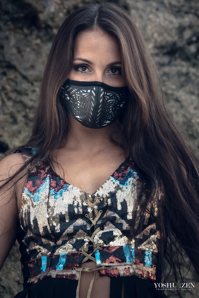 May include: A woman with long brown hair wearing a black face mask with a geometric design. She is wearing a sequined top with a geometric pattern in shades of blue, red, and gold. The top has a lace-up front.