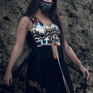 May include: A woman wearing a black, sequined, and fringed top with a black face mask. The top has a gold and blue geometric pattern and a lace-up front. The woman is standing in front of a rocky background.