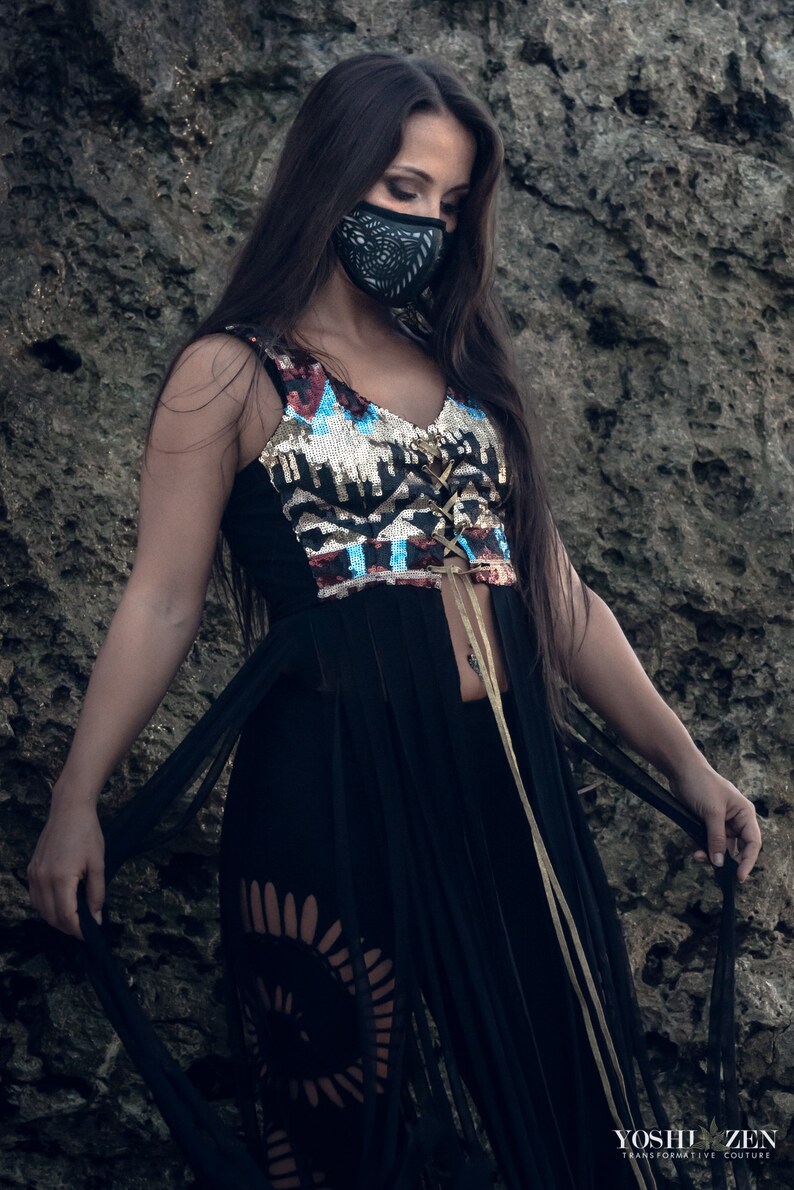 May include: A woman wearing a black, sequined, and fringed top with a black face mask. The top has a gold and blue geometric pattern and a lace-up front. The woman is standing in front of a rocky background.