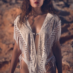 May include: A woman wearing a white crochet top with fringe detailing. The top has a deep V-neckline and a delicate lace pattern. The woman is standing in front of a rocky background.