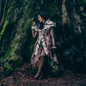 May include: A woman wearing a long, brown and white fur-trimmed coat stands in front of a large tree trunk. The coat has a sequined design and a long, flowing skirt. The woman is wearing brown boots and has long, dark hair.