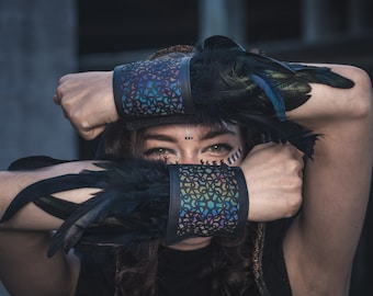 May include: A person wearing black feather wrist cuffs with a geometric pattern. The cuffs are iridescent and have a black leather base.