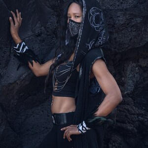 May include: A person wearing a black hooded top with silver embellishments, a black face mask with a geometric design, and black pants with cutouts. The person is standing in front of a dark gray textured wall. The text "YOSHI ZEN" is visible in the lower right corner of the image.