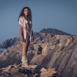 May include: A woman wearing a white crochet top and white bikini bottoms stands on a rocky cliff overlooking the ocean. She is wearing white boots and has long brown hair.