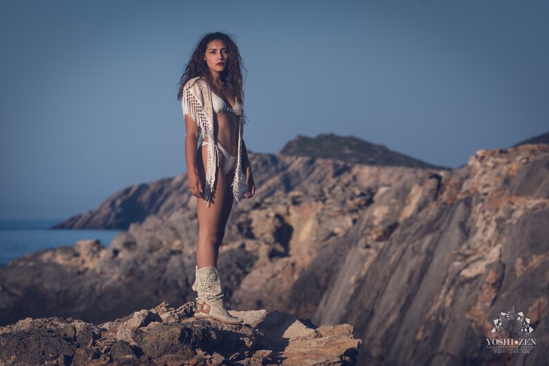 May include: A woman wearing a white crochet top and white bikini bottoms stands on a rocky cliff overlooking the ocean. She is wearing white boots and has long brown hair.