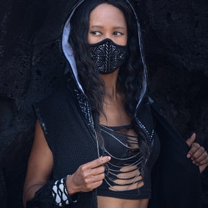 May include: A person wearing a black hooded jacket with a white trim and a black and white geometric face mask. The person is wearing a black top with a silver chain necklace and a black belt with a silver buckle. The person has a belly button ring.