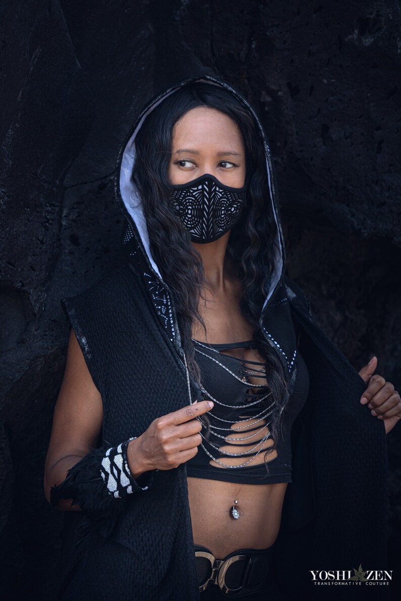 May include: A person wearing a black hooded jacket with a white trim and a black and white geometric face mask. The person is wearing a black top with a silver chain necklace and a black belt with a silver buckle. The person has a belly button ring.