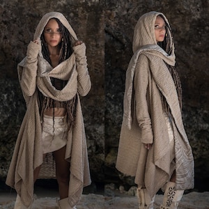 May include: A beige hooded cardigan with a long, flowing design. The cardigan has a textured fabric and a unique asymmetrical hemline. The model is wearing the cardigan with a white crop top and a white skirt.