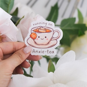 Könnte beinhalten: Weißer Aufkleber mit einer Cartoon-Illustration einer Teetasse mit einem Teebeutel, auf dem "anxie-tea" steht. Der Text "running on" befindet sich über der Teetasse und der Text "anxie-tea" unter der Teetasse.