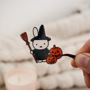 Könnte beinhalten: Ein Halloween-Sticker mit einem Cartoon-Hasen als Hexe, der einen Besen hält und von zwei geschnitzten Kürbissen begleitet wird. Der Hase trägt einen schwarzen Hexenhut mit Sternen.