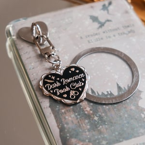 Pode incluir: Um porta-chaves prateado com um pendente em forma de coração. O pendente é preto com contorno branco e apresenta o texto "Dark Romance Book Club". Um marcador de livros prateado em forma de anel também é visível.