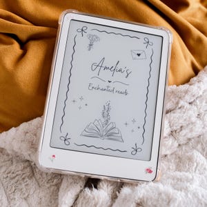 Könnte beinhalten: Ein weißer E-Reader mit einer transparenten Schutzhülle. Auf dem Bildschirm steht "Amelia's Enchanted Reads" mit floralen Illustrationen. Das Gerät ist mit kleinen floralen Akzenten verziert und liegt auf einer weichen, strukturierten Oberfläche.